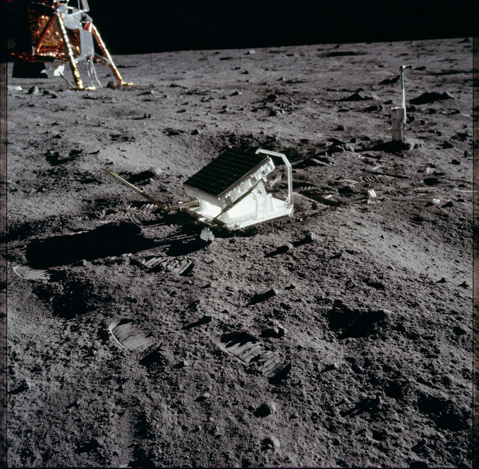 Panel cuadrado de prismas apoyado sobre el suelo gris de la Luna, con el módulo lunar al fondo.