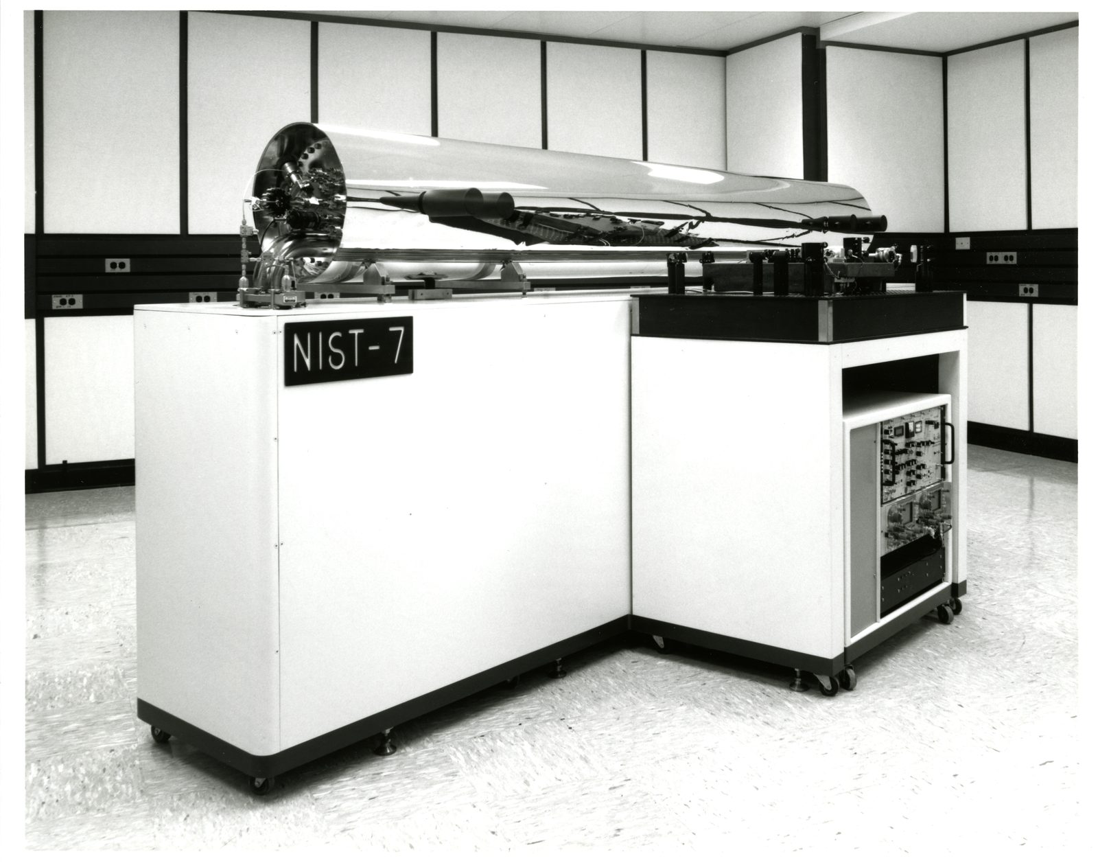 Fotografía en blanco y negro de un reloj atómico de laboratorio: un tubo cilíndrico pulido tendido sobre los gabinetes de instrumentación, con el rótulo NIST-7 en el frente.