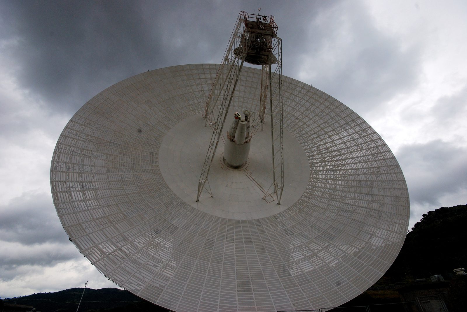 Un enorme plato de antena visto desde abajo, con las cuatro patas que sostienen el subreflector, bajo un cielo gris de tormenta y con un monte al fondo.