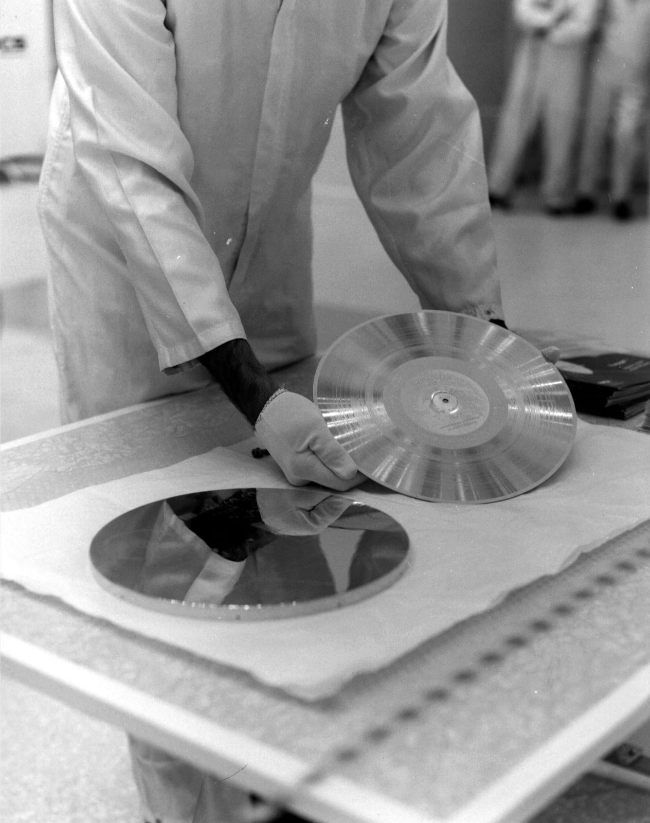 Fotografía en blanco y negro de un técnico de bata blanca y guantes que levanta un disco fonográfico circular sobre una mesa de sala limpia.