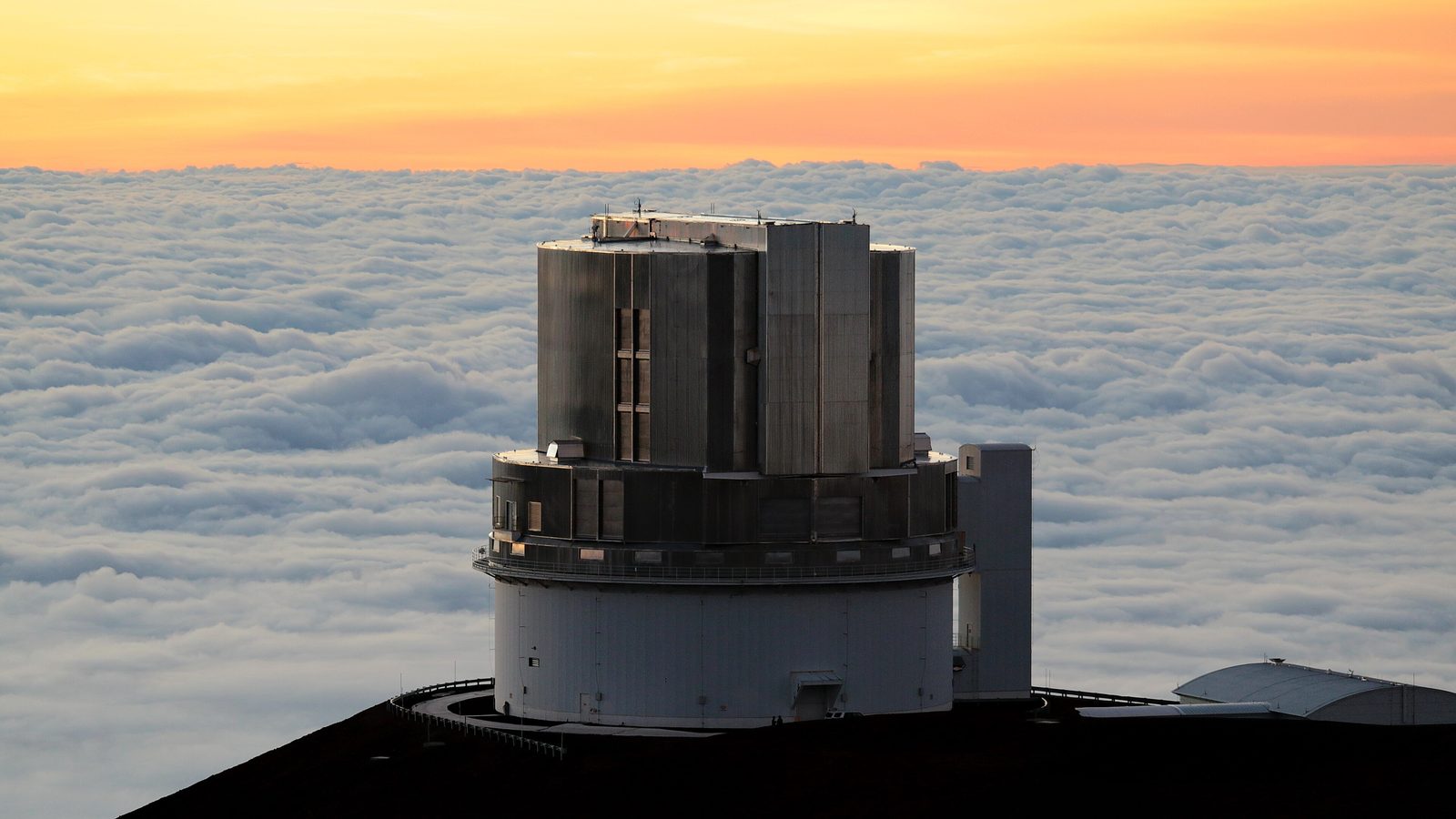 Cúpula cilíndrica de un gran telescopio en la cumbre de una montaña, recortada contra el cielo anaranjado del atardecer por encima de un mar de nubes.