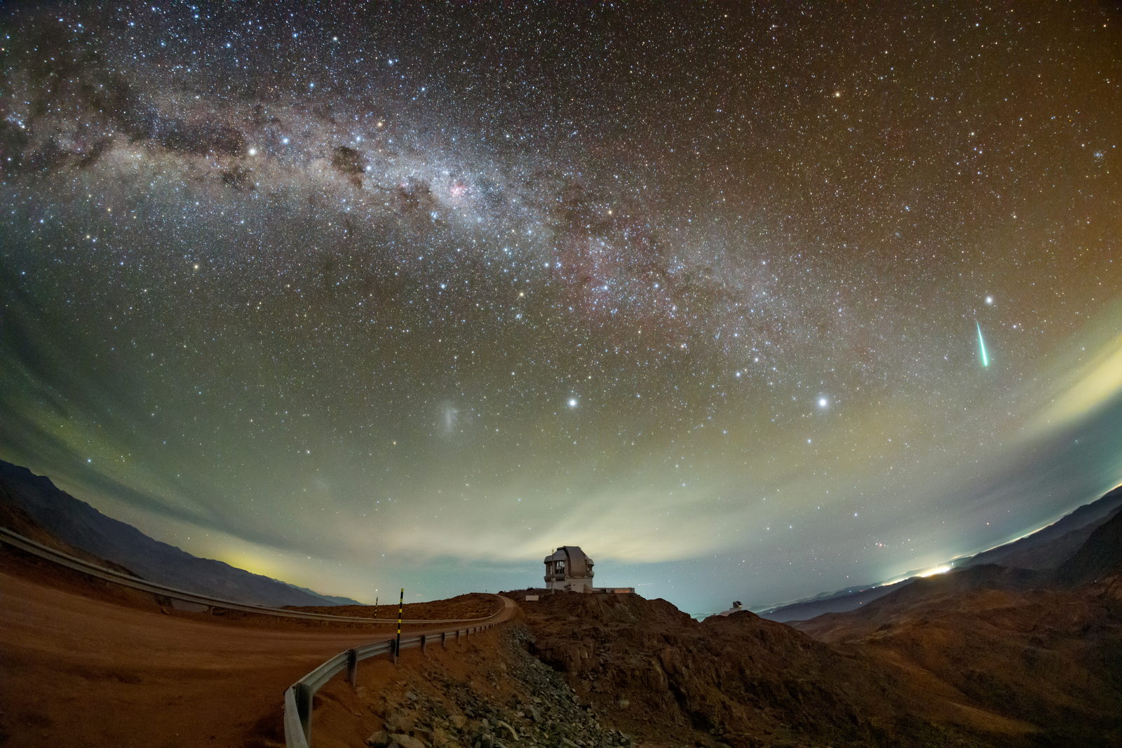 Un meteoro brillante sobre las cúpulas de Cerro Pachón, con las luces de una ciudad lejana en el horizonte y el cielo estrellado encima.