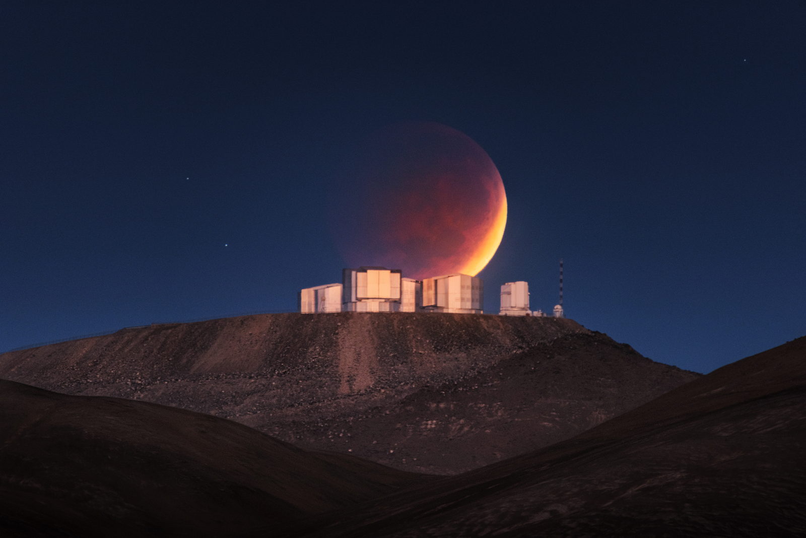 La Luna eclipsada, grande y de color rojo cobrizo, hundiéndose detrás de las cuatro unidades del Telescopio Muy Grande recortadas en lo alto de un cerro, con el cielo del amanecer azul detrás.
