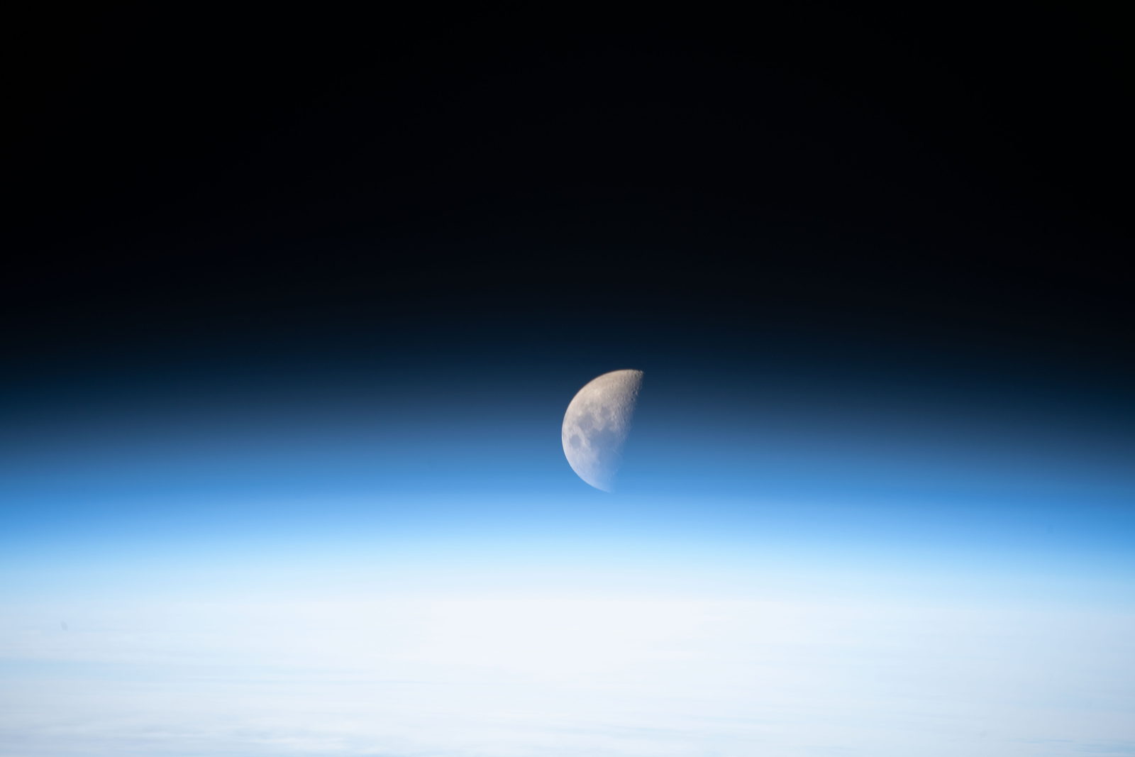 La Luna en cuarto creciente, mitad iluminada y mitad en sombra, sobre el arco azul de la atmósfera terrestre.