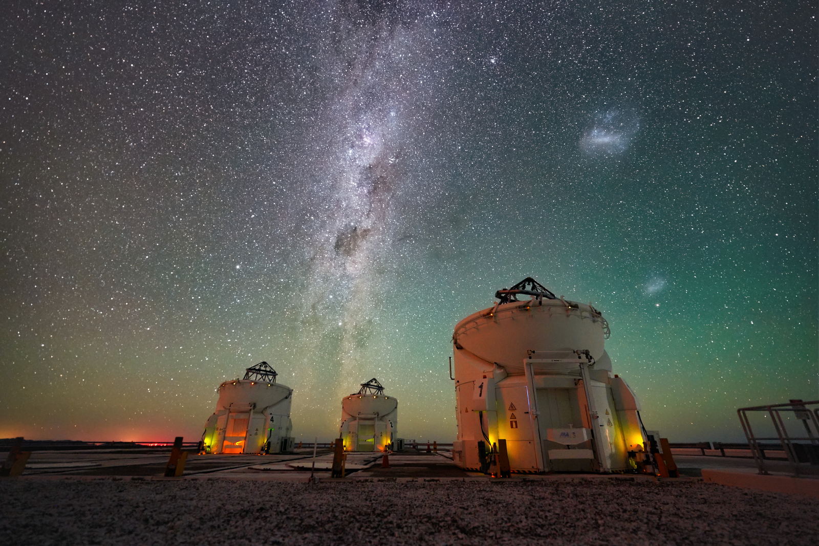 Cielo nocturno con la banda de la Vía Láctea y dos manchas difusas, las Nubes de Magallanes, sobre tres telescopios pequeños en el desierto.