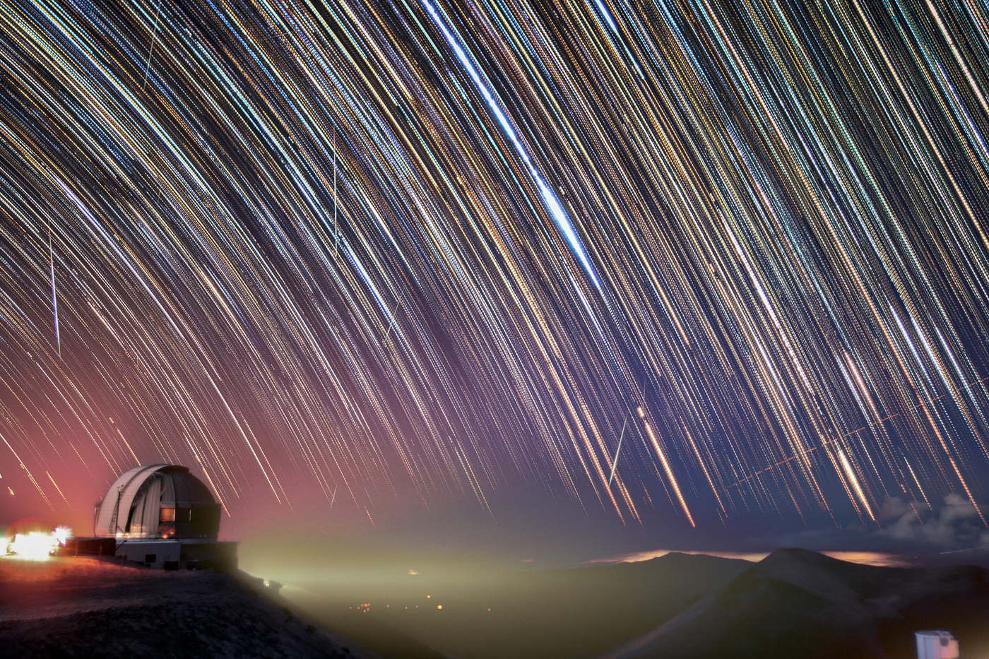 El cielo sobre la cúpula del telescopio Gemini Norte convertido en arcos de colores por una exposición larga; unos pocos trazos rectos los cruzan en diagonal.